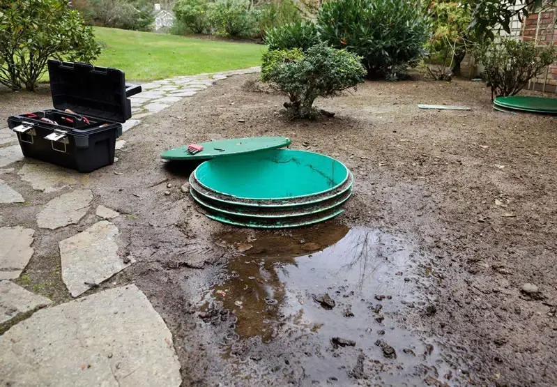 Open septic tank lid with effluent pooled on saturated ground beside a home